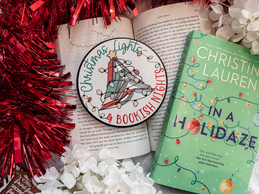 Round shelf sign with a white background. Image of books stacked into a Christmas tree surrounded by lights. The surrounding text reads "Christmas Lights" above in green, with "& Bookish Nights" below in red. © FireDrake Artistry 2025