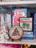 Load image into Gallery viewer, Round shelf sign with a white background. Image of books stacked into a Christmas tree surrounded by lights. The surrounding text reads "Christmas Lights" above in green, with "& Bookish Nights" below in red. © FireDrake Artistry 2025
