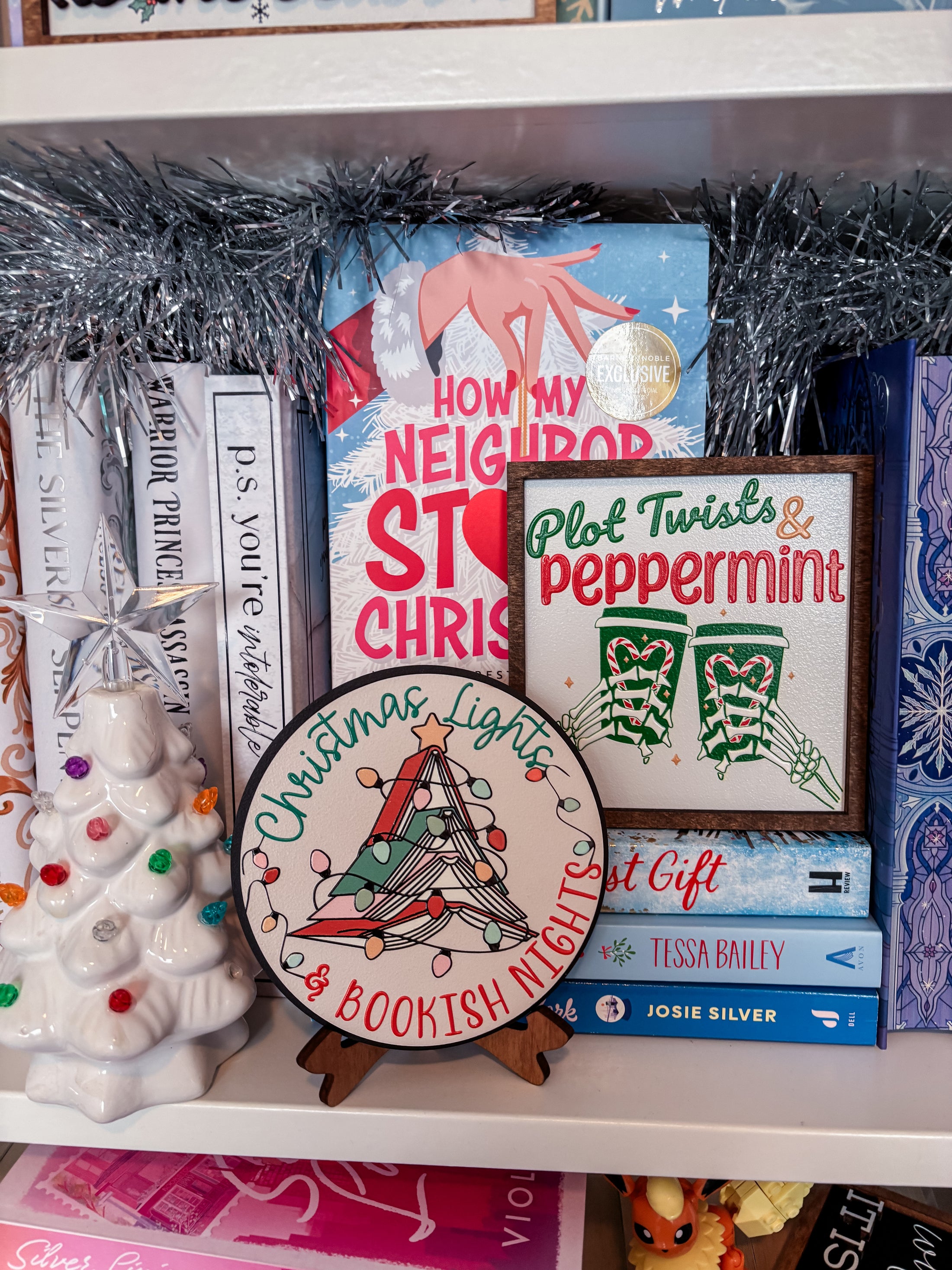Round shelf sign with a white background. Image of books stacked into a Christmas tree surrounded by lights. The surrounding text reads "Christmas Lights" above in green, with "& Bookish Nights" below in red. © FireDrake Artistry 2025