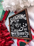 Load image into Gallery viewer, Wooden shelf sign with a black background and woodgrain frame.  A white sleigh with presents, surrounded by white text reading  'Sleighing My TBR' and "Winter Reader Co. EST 2025." © FireDrake Artistry 2025
