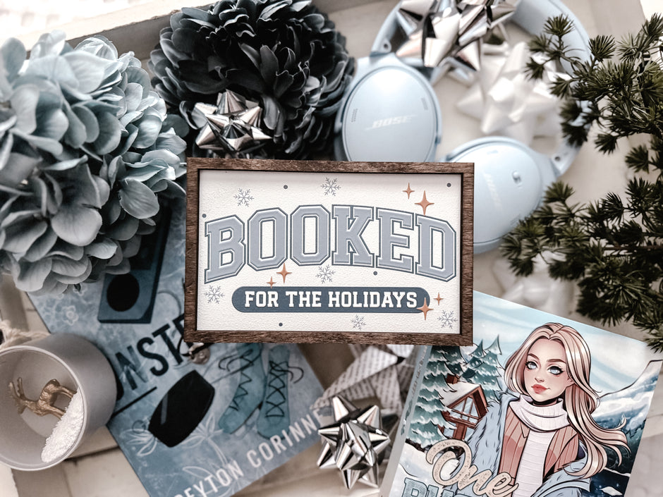 Decorative sign with a wooden frame that reads 'Booked for the Holidays' in blue on a white background. © FireDrake Artistry 2025.