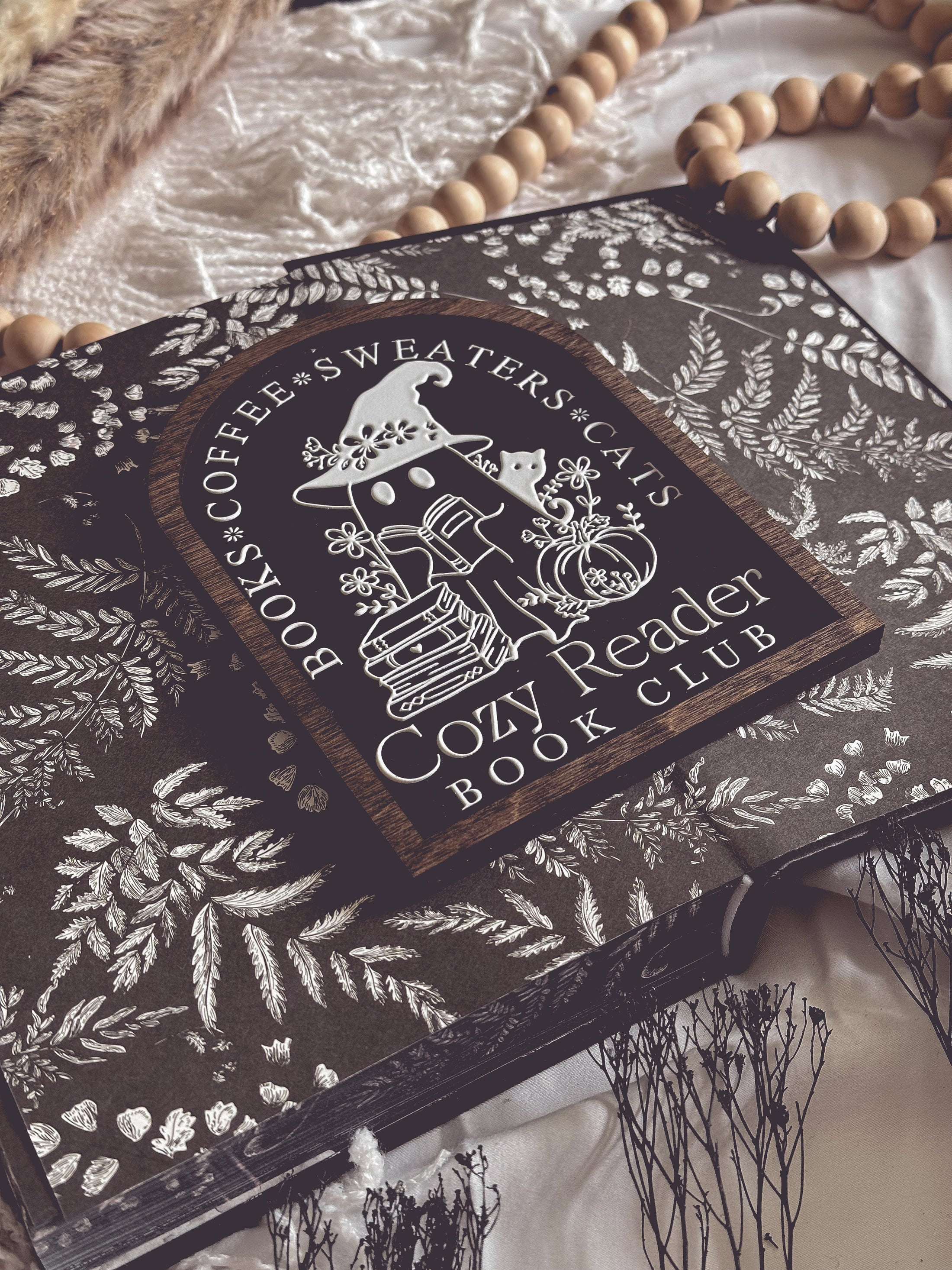 Cozy Reader Ghosty Sign. Black background with a dark wood border with a white design engraved. 
Featuring a ghost reading a book and wearing a witch hat, surrounded by flowers and a pumpkin. Text above reads “Books. Coffee. Sweaters. Cats.” While the text below reads “Cozy Reader Book Club.” 
