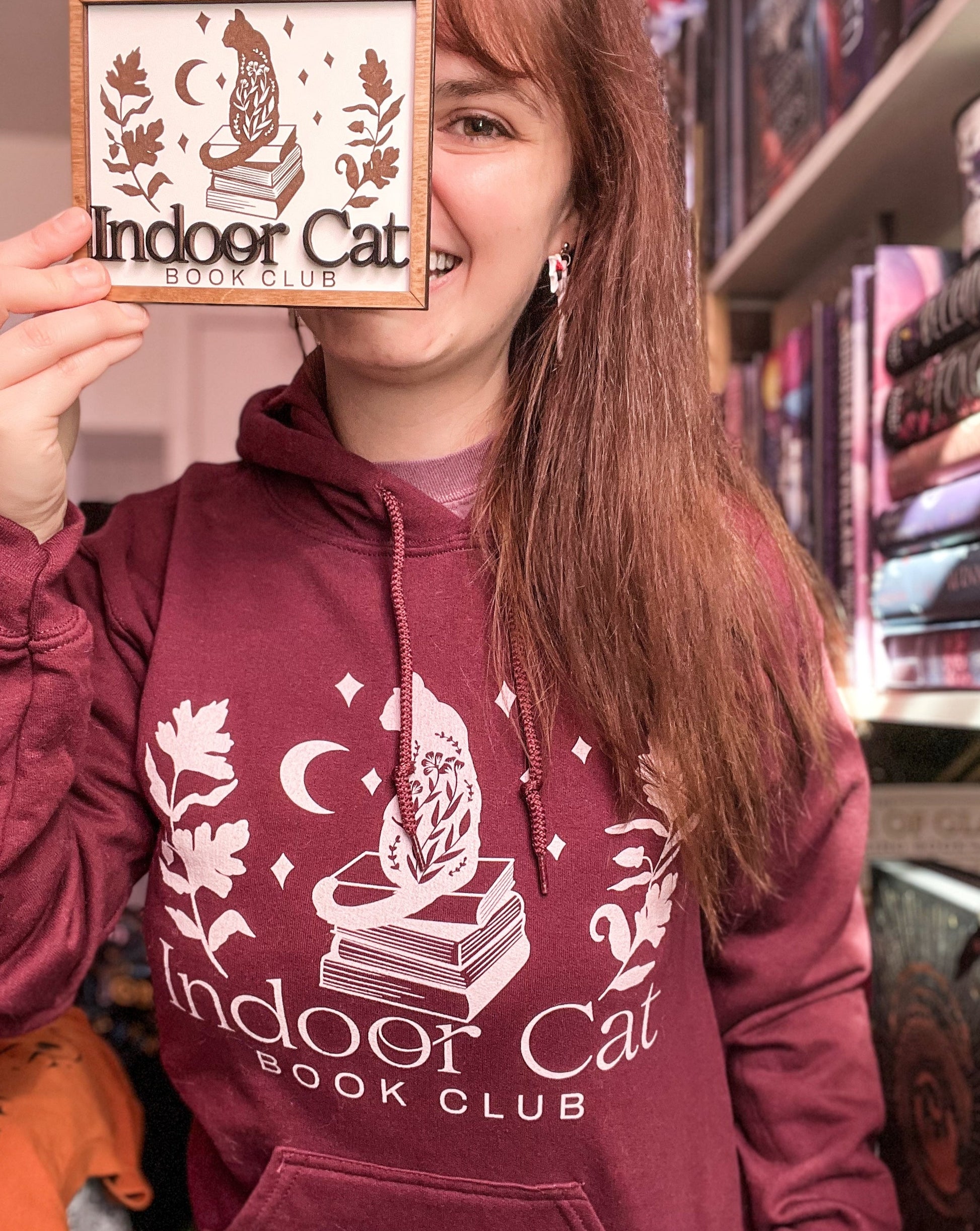 Woman next to bookshelf wearing indoor cat hoodie in maroon with white design