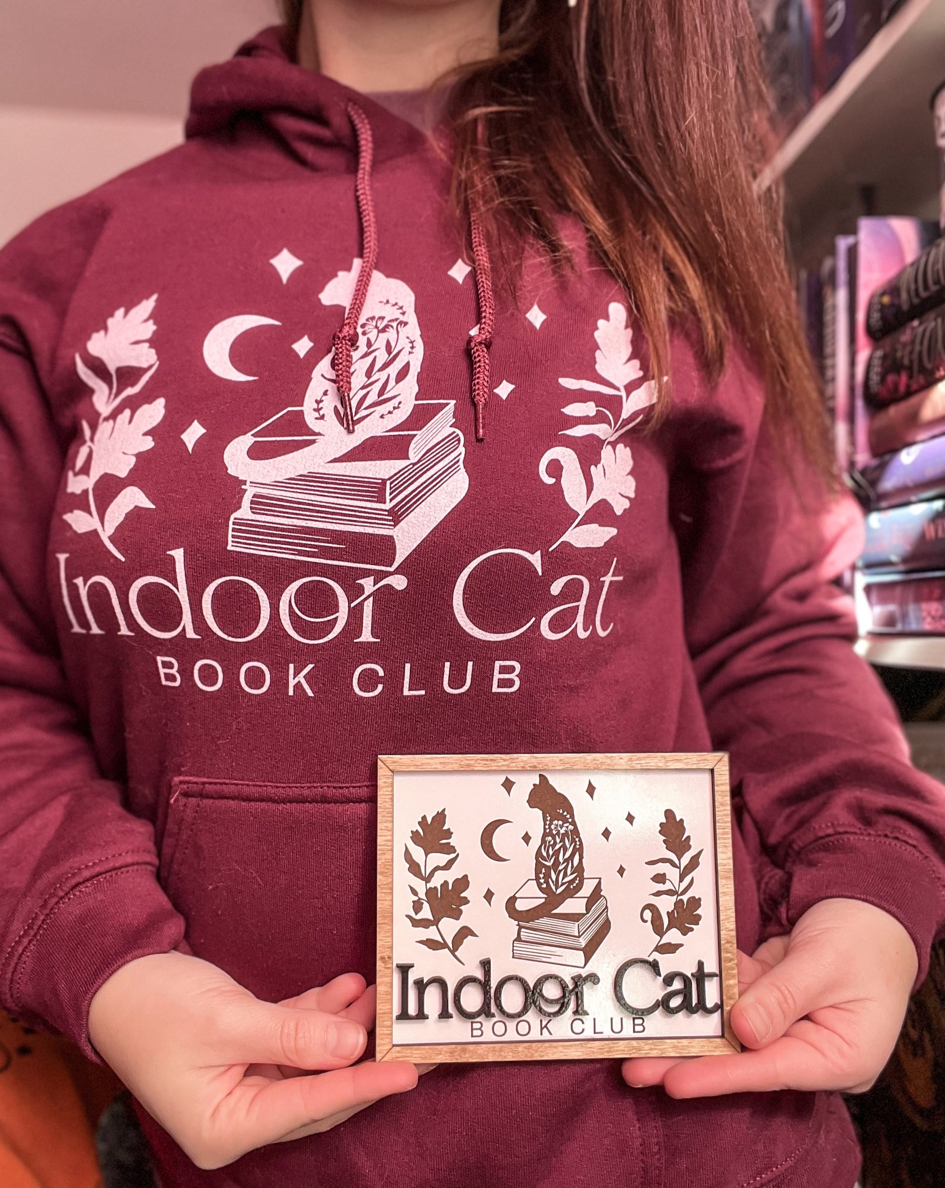 Woman next to bookshelf wearing indoor cat hoodie in maroon with white design