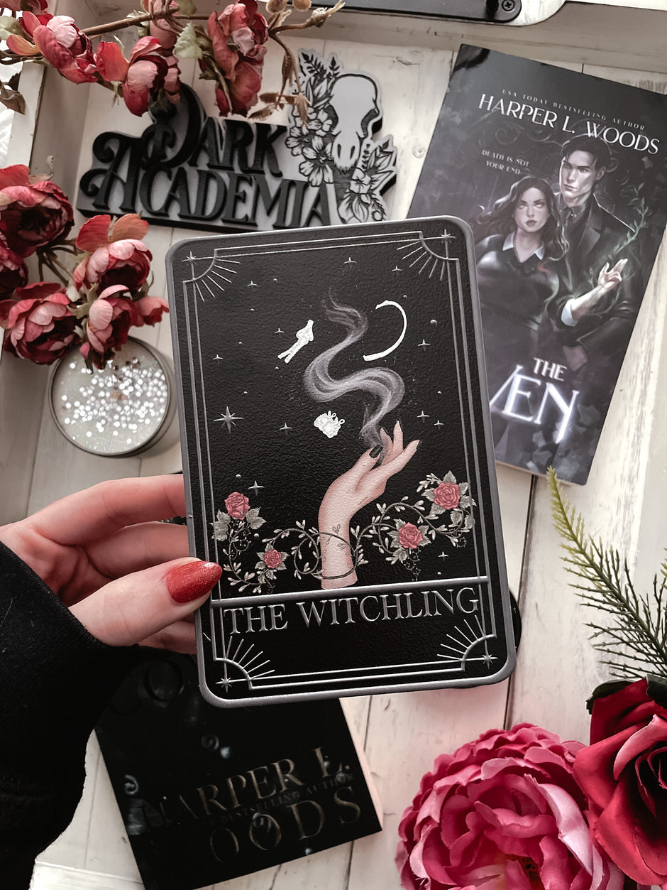 A hand holding a Tarot Card titled "The Witching" with a magical hand printed on it, on top of a wooden tray with a book, shelf sign, and flowers in the background. 