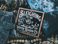 Load image into Gallery viewer, Wooden shelf sign with a black background and woodgrain frame.  A white sleigh with presents, surrounded by white text reading  'Sleighing My TBR' and "Winter Reader Co. EST 2025." © FireDrake Artistry 2025
