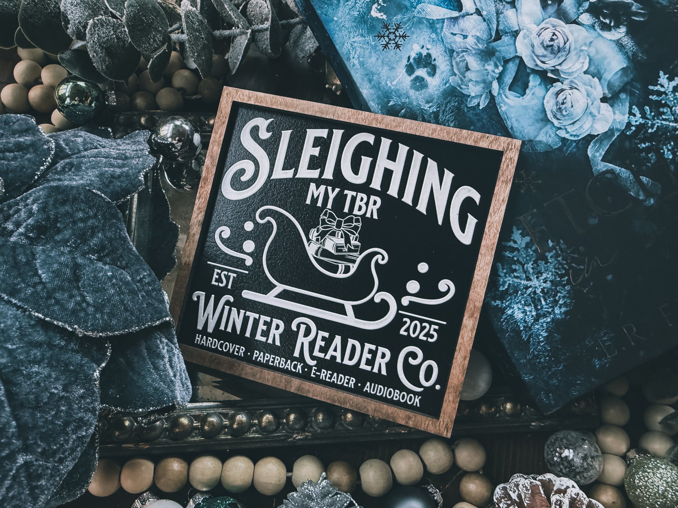 Wooden shelf sign with a black background and woodgrain frame.  A white sleigh with presents, surrounded by white text reading  'Sleighing My TBR' and "Winter Reader Co. EST 2025." © FireDrake Artistry 2025
