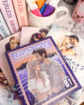 Load image into Gallery viewer, A postage stamp featuring artwork of a romantic scene, on top of a stack of books with a book stack and mug with colorful highlighters in the background. 
