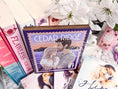 Load image into Gallery viewer, A postage stamp featuring artwork of a romantic scene, on top of a stack of books with a book stack and mug with flowers in the background. 
