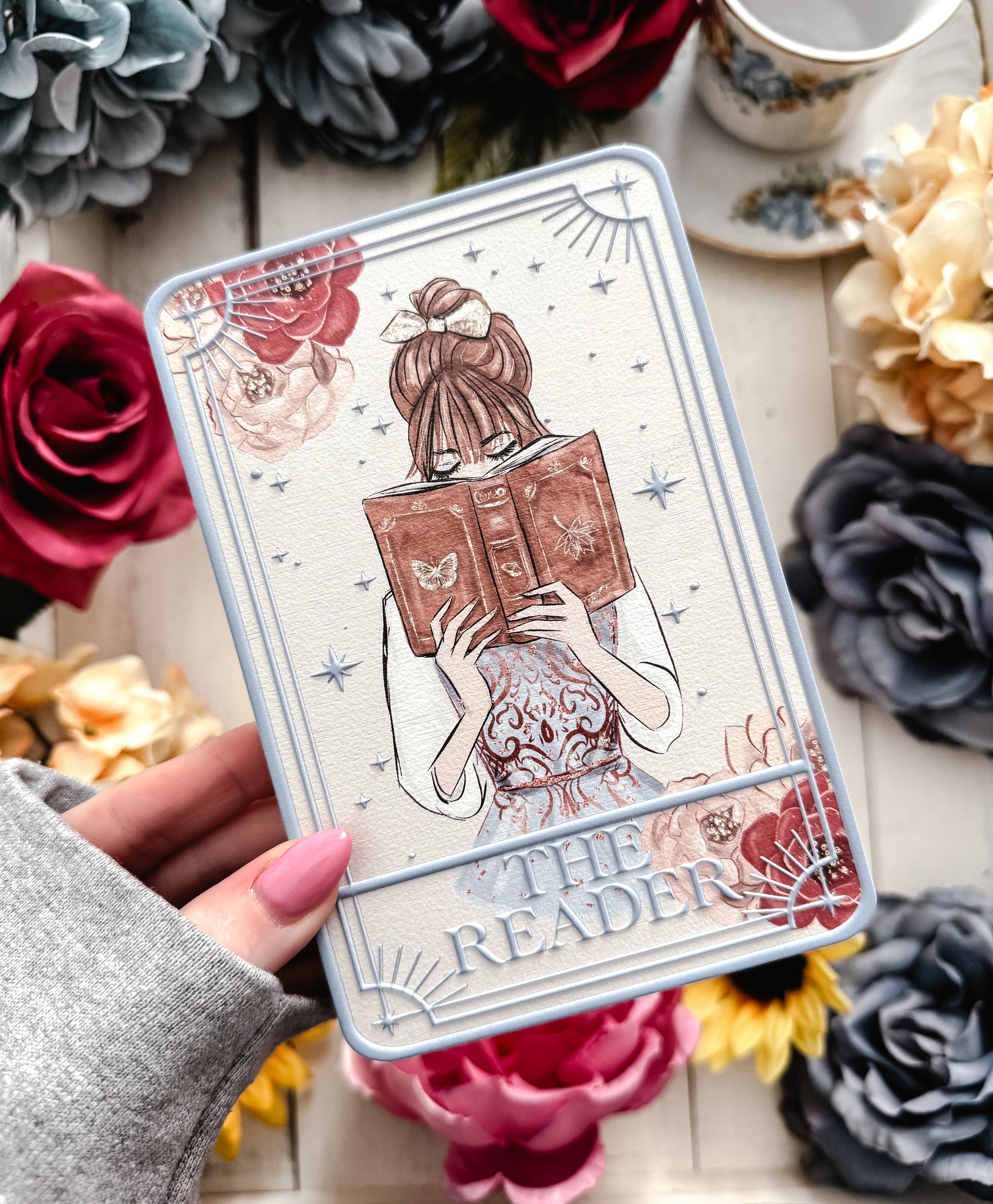 Tarot card title “The Reader” with a girl reading an open book being held in a hand with flowers and other decorative items in the background. 