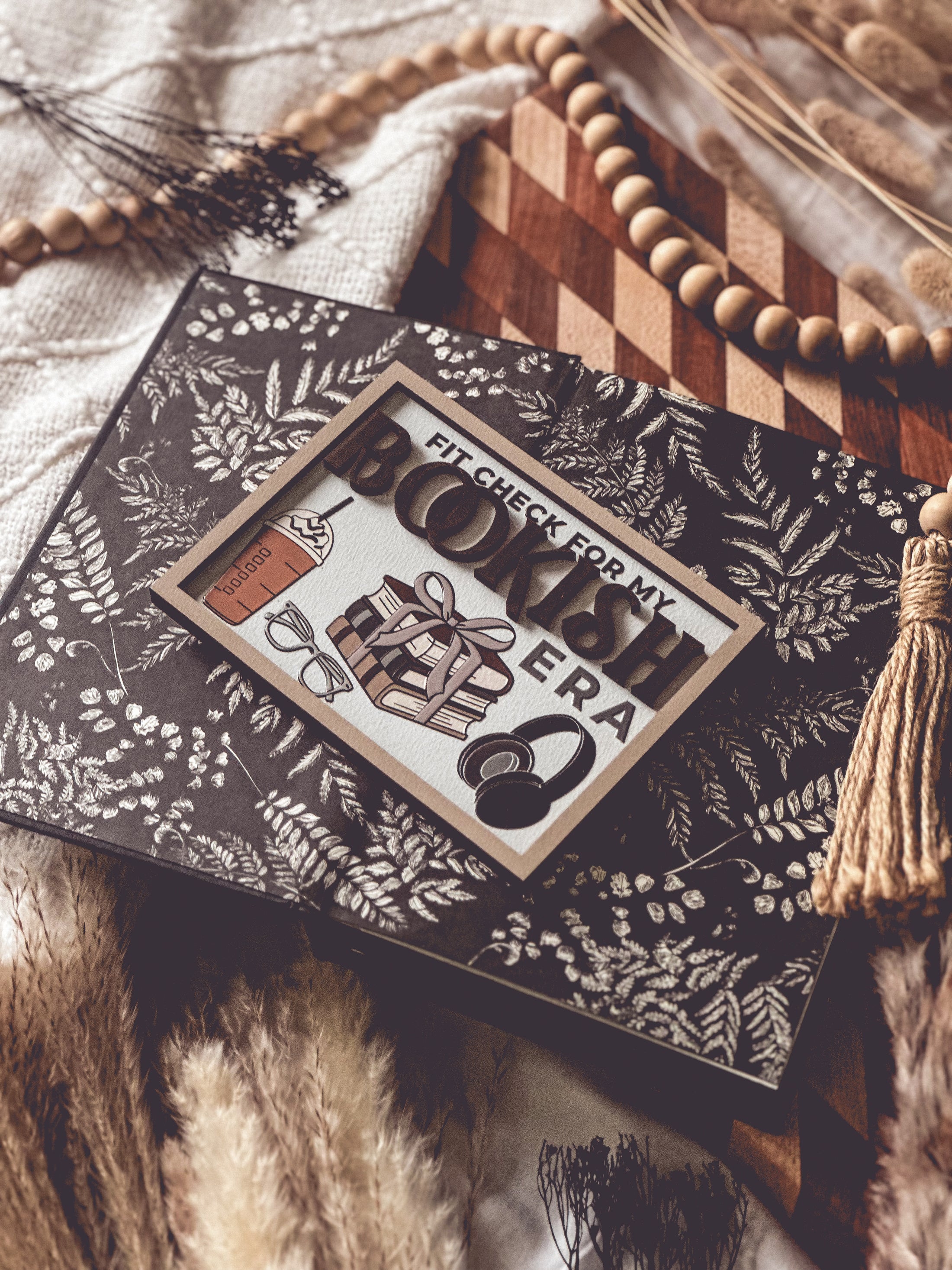 Wooden sign with a white background and tan border. Text reads "Fit Check For My Bookish Era" with surrounding graphics of a latte, glasses, a stack of books, and headphones. © FireDrake Artistry