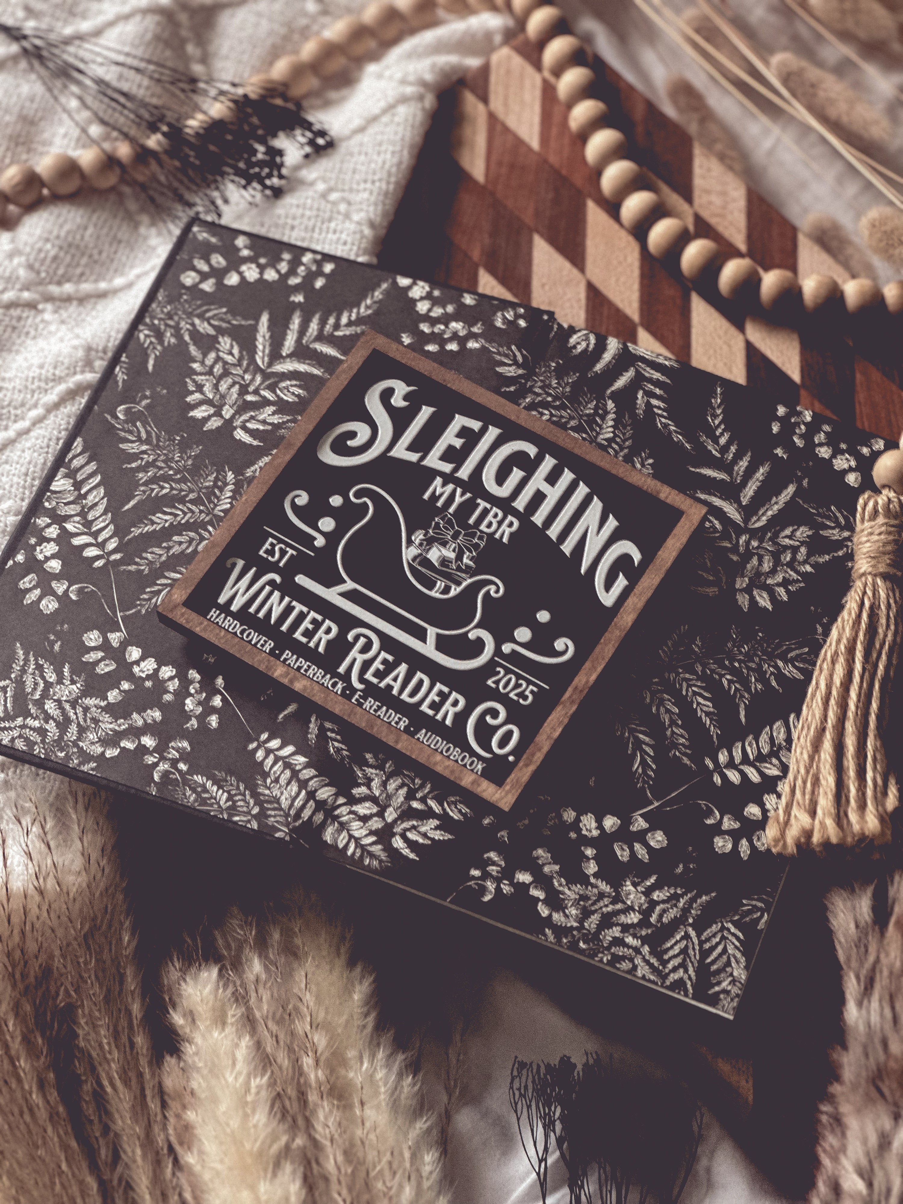 Wooden shelf sign with a black background and woodgrain frame.  A white sleigh with presents, surrounded by white text reading  'Sleighing My TBR' and "Winter Reader Co. EST 2025." © FireDrake Artistry 2025