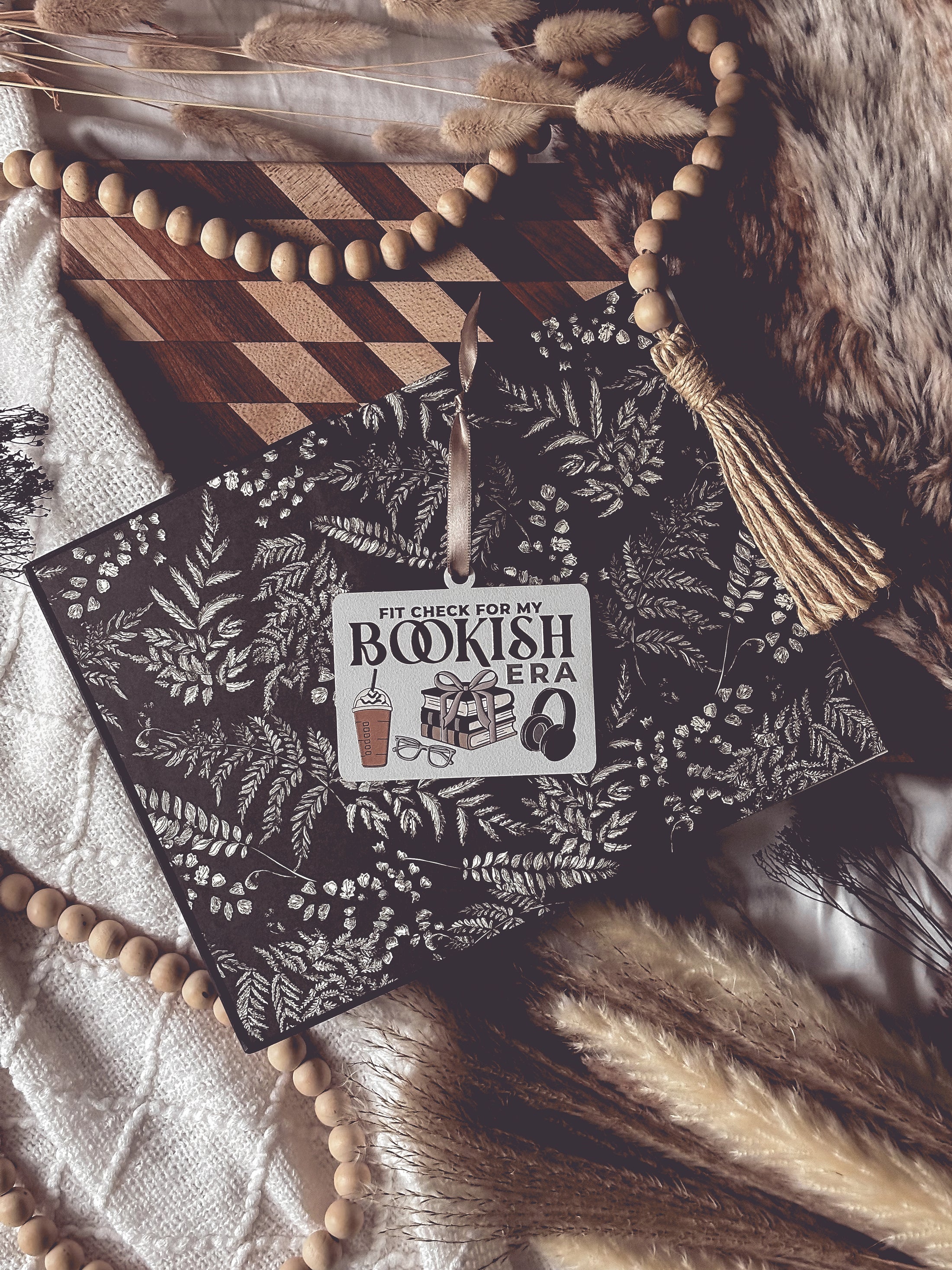 Wooden ornament with a white background and tan ribbon. Text reads "Fit Check For My Bookish Era" with surrounding graphics of a latte, glasses, a stack of books, and headphones. © FireDrake Artistry