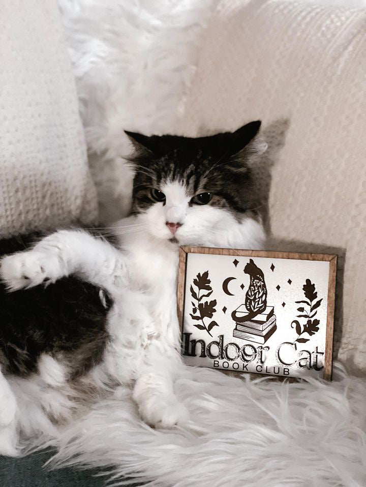 Indoor Cat Book Club Sign. White wooden sign with a wood frame. Design featuring a floral cat sitting atop a pile of books flanked by flowers, a moon, and some stars. Text below reads "Indoor Cat Book Club". Featuring a grey and white cat. 2023 FireDrake Artistry®