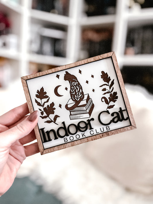 Indoor Cat Book Club Sign. White wooden sign with a wood frame. Design featuring a floral cat sitting atop a pile of books flanked by flowers, a moon, and some stars. Text below reads "Indoor Cat Book Club". 2023 FireDrake Artistry®