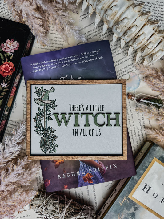 A white shelf sign with a wood grain border, featuring text that reads “There’s a little witch in all of us” and a graphic of a witch hat on a broomstick surrounded by flowers. ©️FireDrake Artistry®️ 2025