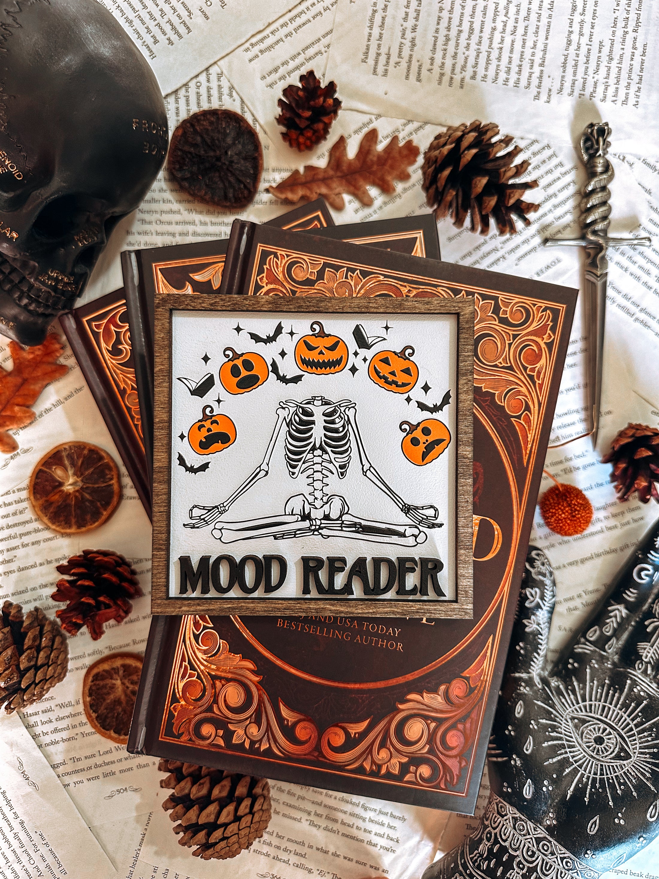 Mood Reader Sign. A square decorative sign made of wood with a wood-stained frame. UV printed skeleton in a zen position, surrounded by pumpkins with multiple faces, with the text 'MOOD READER' across the center. FireDrake Artistry 2025