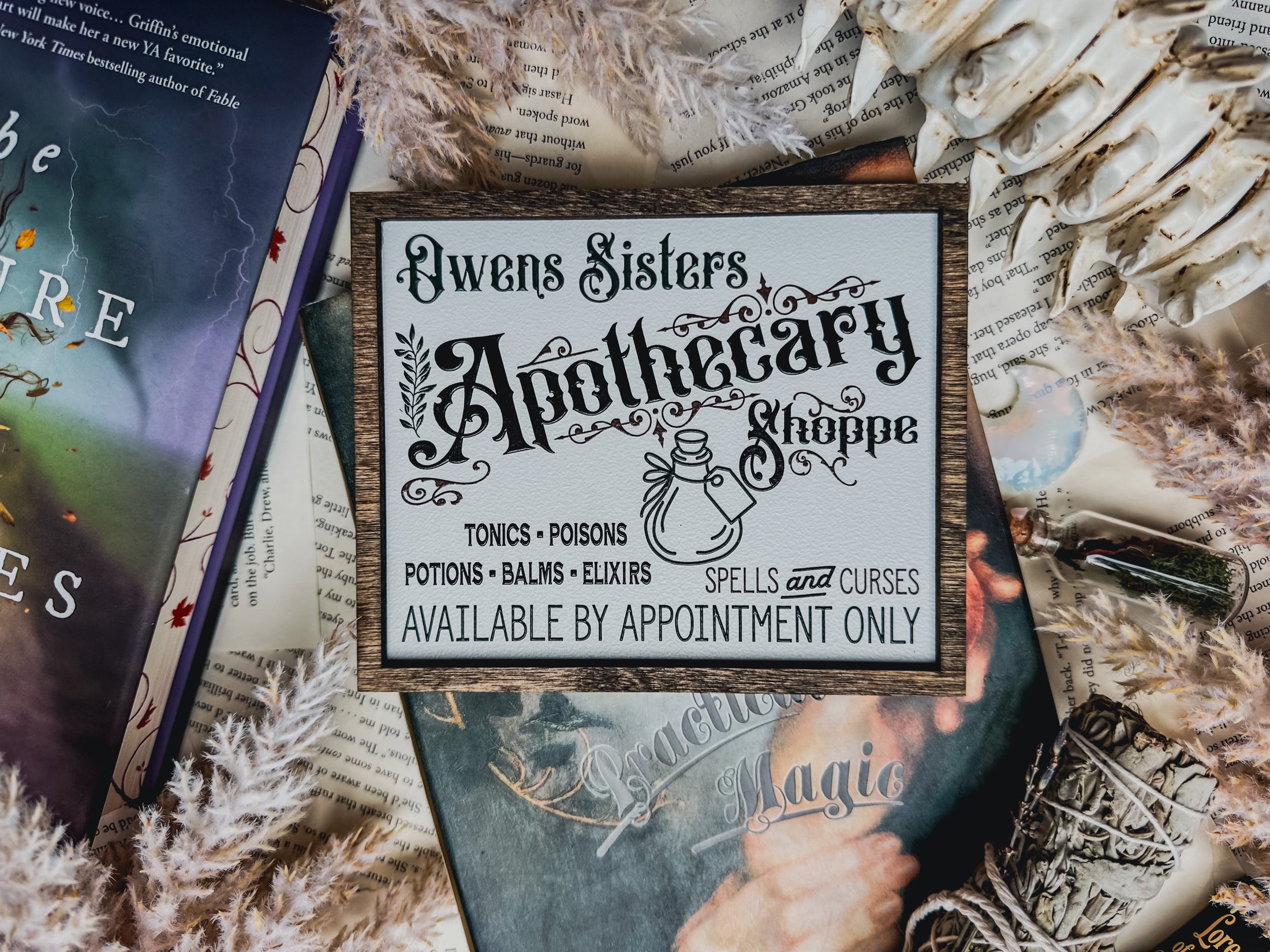 White shelf sign with a wood grain border. Featuring text thy reads “Owens Sisters’ Apothocary Shoppe” with a list of ingredients, a note for appointment only, and a tea pot. ©️FireDrake Artistry®️ 2025