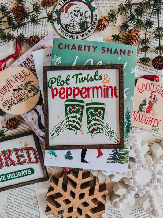 Decorative sign with a dark wood background, 'Plot Twists & Peppermint' text, with skeleton hands holding hot chocolate, on a white background. © FireDrake Artistry 2025