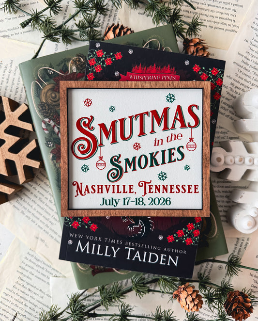 Woods sign with "Smutmas in the Smokies" and other event information printed on it, on top of red and green books. Book pages in the background with other holiday decor. 