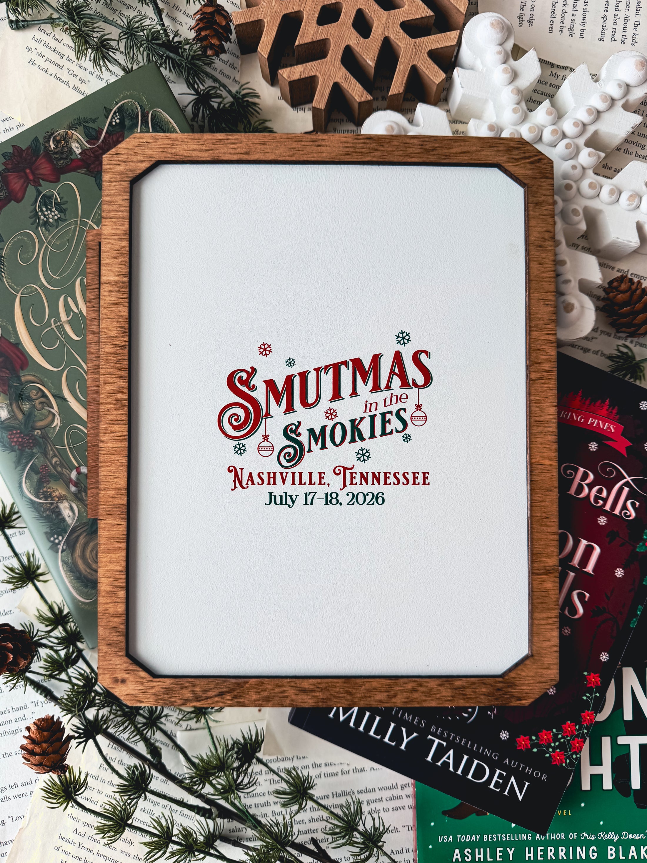 Wooden sign with frame and white base over books and wooden snowflakes. "Smutmas in the Smokies" event information printed on the board.