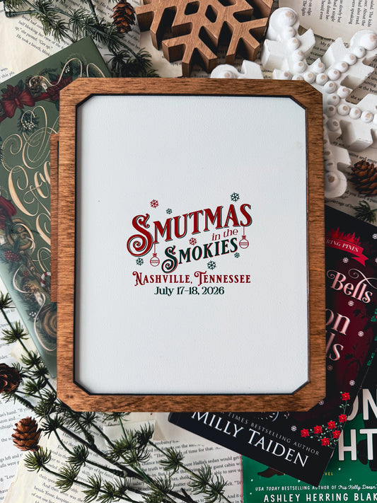 Wooden sign with frame and white base over books and wooden snowflakes. "Smutmas in the Smokies" event information printed on the board.